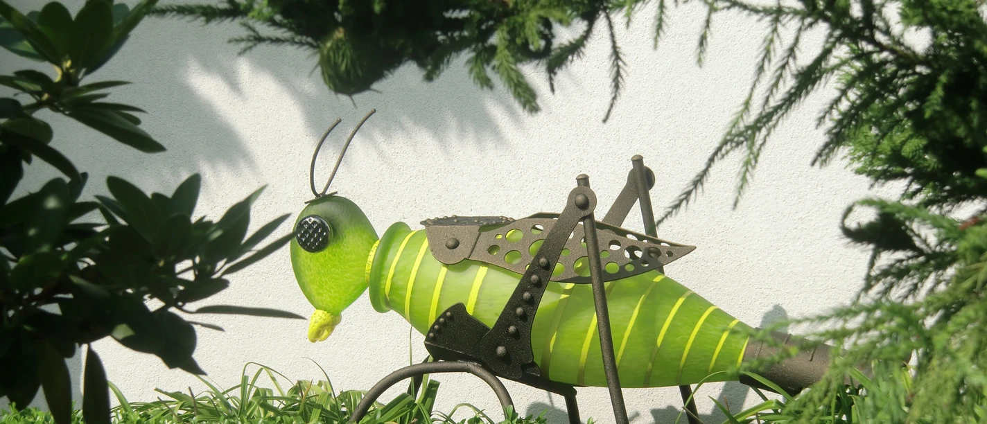 Galerie Spiegel - Skulptur Grille Grüne Grashüpfer-Skulptur aus Metall, umgeben von dichten Sträuchern und Blättern im Sonnenlicht.