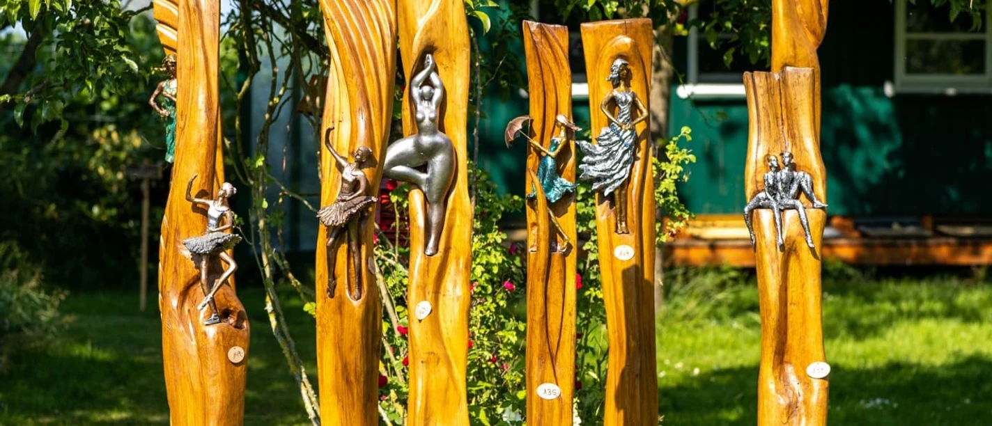 Handwerkermarkt_Christa Staffa.jpg Fünf kunstvoll geschnitzte Holzstämme mit metallischen Verzierungen stehen in einem Garten.