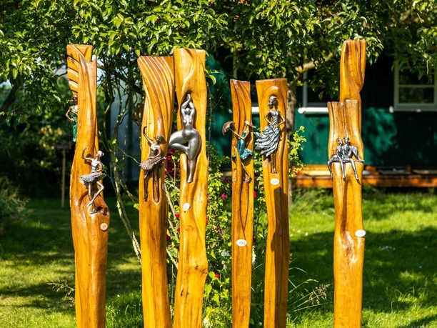 Handwerkermarkt_Christa Staffa.jpg Fünf kunstvoll geschnitzte Holzstämme mit metallischen Verzierungen stehen in einem Garten.