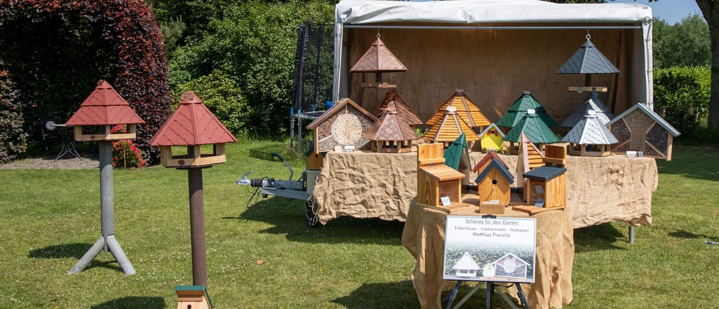 Futter Häuser_Handwerkermarkt-Tange_Christa Staffa.jpg Das Bild zeigt einen Stand auf einem Handwerkermarkt mit kunstvoll gefertigten Vogelhäusern in vielfältigen Farben und Formen. Im Hintergrund steht ein großer Pavillon, unter dem die Vogelhäuser auf einem Tisch präsentiert werden. Rundherum befindet sich eine grüne Rasenfläche und dichte Baumreihen, die eine angenehme, ländliche Atmosphäre erzeugen.