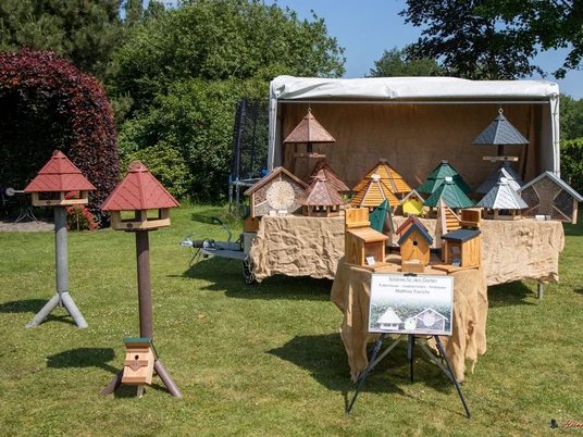 Futter Häuser_Handwerkermarkt-Tange_Christa Staffa.jpg Das Bild zeigt einen Stand auf einem Handwerkermarkt mit kunstvoll gefertigten Vogelhäusern in vielfältigen Farben und Formen. Im Hintergrund steht ein großer Pavillon, unter dem die Vogelhäuser auf einem Tisch präsentiert werden. Rundherum befindet sich eine grüne Rasenfläche und dichte Baumreihen, die eine angenehme, ländliche Atmosphäre erzeugen.