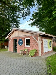 HOF.echt Hofladen Fam. Theissing in Salzbergen ©Emsland Tourismus GmbH (1).jpg Kleines Backsteingebäude mit Holzgiebel und HOF.echt-Schild, umgeben von Bäumen und Grünfläche.