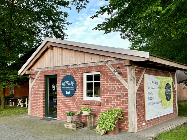 HOF.echt Hofladen Fam. Theissing in Salzbergen ©Emsland Tourismus GmbH (1).jpg Kleines Backsteingebäude mit Holzgiebel und HOF.echt-Schild, umgeben von Bäumen und Grünfläche.