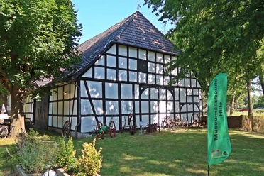 Weiße Fachwerkscheune mit dunklen Holzbalken und ziegelgedecktem Dach in grüner Umgebung.