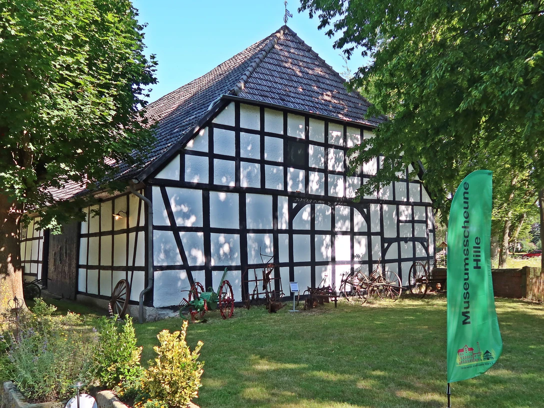 Weiße Fachwerkscheune mit dunklen Holzbalken und ziegelgedecktem Dach in grüner Umgebung.