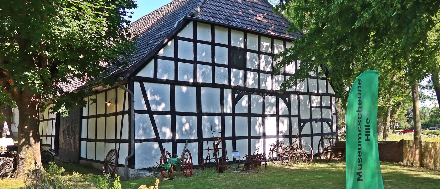 Museumsscheune Hille - Außenansicht Weiße Fachwerkscheune mit dunklen Holzbalken und ziegelgedecktem Dach in grüner Umgebung.
