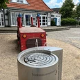 Trinkwasser-Brunnen Roter Holztraktor mit Fontäne im Vordergrund, stehend auf gepflastertem Platz vor weißem Gebäude.