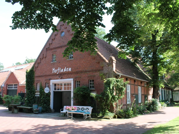 Schroeter & Wielage, Bawinkel - Außenansicht Hofladen ©Emsland Tourismus GmbH (4).JPG Backsteingebäude mit Schriftzug Hofladen, umgeben von Bäumen, Blumen und Sitzbank im Eingangsbereich.