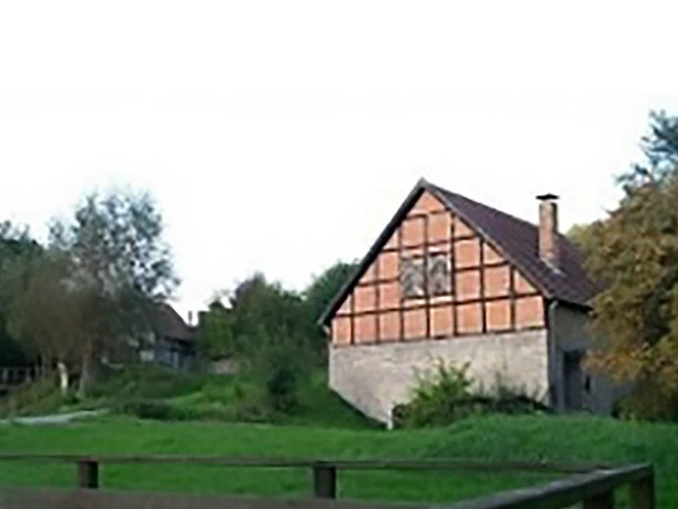 Fachwerkhaus mit rotem Ziegeldach in grüner Landschaft, umgeben von Bäumen und Zaun im Vordergrund.