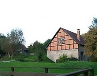 Bad Driburg_Schoepfmuehle_Dringenberg Fachwerkhaus mit rotem Ziegeldach in grüner Landschaft, umgeben von Bäumen und Zaun im Vordergrund.