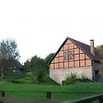 Bad Driburg_Schoepfmuehle_Dringenberg Fachwerkhaus mit rotem Ziegeldach in grüner Landschaft, umgeben von Bäumen und Zaun im Vordergrund.