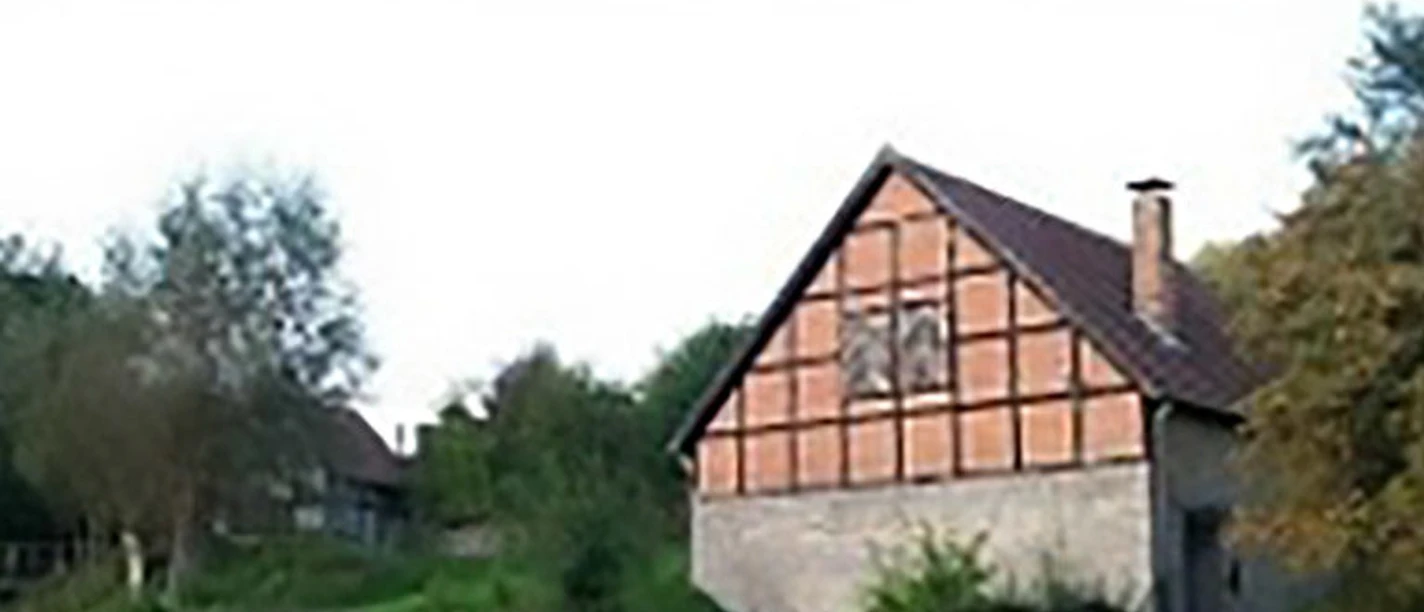 Bad Driburg_Schoepfmuehle_Dringenberg Fachwerkhaus mit rotem Ziegeldach in grüner Landschaft, umgeben von Bäumen und Zaun im Vordergrund.