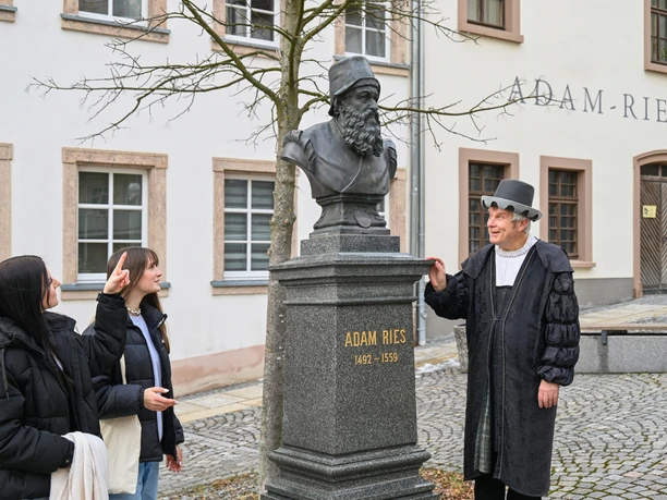 adam-ries-museum-fotoshooting-foto-annett-flaemig-stadt-annaberg-2.jpg Zwei junge Frauen und ein Mann im historischen Kostüm betrachten eine Adam-Ries-Büste.