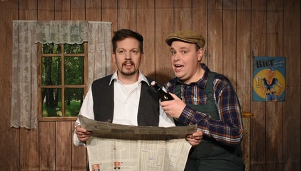Pressefoto-Landei-gesucht ©ZIMMER.THEATER.png Zwei Männer in Bauernkleidung stehen in rustikalem Raum, einer liest Zeitung, der andere hält Bier.