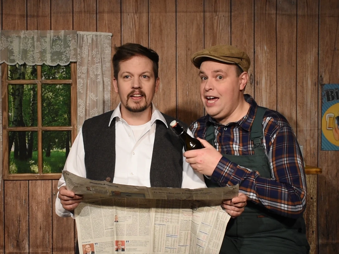 Pressefoto-Landei-gesucht ©ZIMMER.THEATER.png Zwei Männer in Bauernkleidung stehen in rustikalem Raum, einer liest Zeitung, der andere hält Bier.