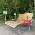Holzliegebank am Waldrand mit roter Decke, Wasserflasche und Hinweisschildern auf Naturlehrpfad.