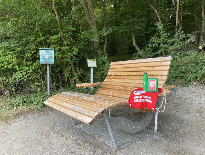 Rastplatz Schmecken NEU Holzliegebank am Waldrand mit roter Decke, Wasserflasche und Hinweisschildern auf Naturlehrpfad.