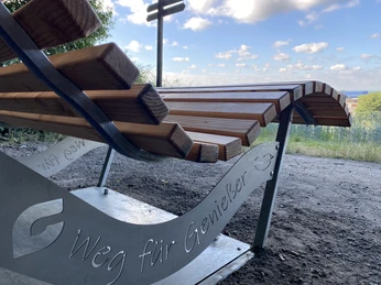 Rastplatz Schmecken NEU Schriftzug Holzliegebank am Waldrand mit der Gravur "Weg für Genießer", umgeben von Bäumen und weitem Himmelblick.