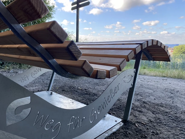 Rastplatz Schmecken NEU Schriftzug Holzliegebank am Waldrand mit der Gravur "Weg für Genießer", umgeben von Bäumen und weitem Himmelblick.