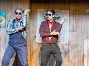 Pressefoto-Landei-gesucht-1 ©ZIMMER.THEATER.JPG Zwei Männer in Bühnenkostümen tanzen vor hölzerner Wandkulisse mit witziger Gestik und Sonnenbrillen.