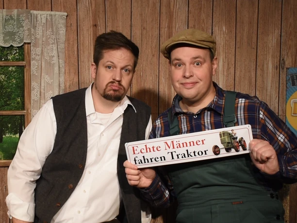 Pressefoto-Landei-gesucht-2 ©ZIMMER.THEATER.png Zwei Männer in ländlicher Kleidung posieren in einer Holzhütte mit einem Schild „Echte Männer fahren Traktor“.