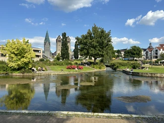 Westliches Paderquellgebiet Grüne Parklandschaft mit Fluss und Blick auf historische Gebäude bei blauem Himmel in Paderborn.