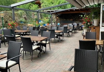 Elliniko.png Das Bild zeigt die Außenterrasse des Restaurants mit mehreren schwarzen Tischen und Stühlen, die auf einem gepflasterten Boden angeordnet sind. Im Hintergrund befindet sich eine überdachte Bar sowie dekorative Pflanzen und Laternen