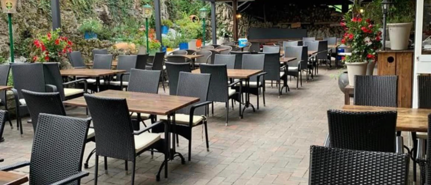 Elliniko.png Das Bild zeigt die Außenterrasse des Restaurants mit mehreren schwarzen Tischen und Stühlen, die auf einem gepflasterten Boden angeordnet sind. Im Hintergrund befindet sich eine überdachte Bar sowie dekorative Pflanzen und Laternen