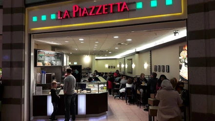 lapiazetta.jpg Das Bild zeigt den Imbiss "La Piazetta". Rechts sind Tische und Stühle zu sehen, während sich auf der linken Seite eine Theke befindet, an der ein Mann eine Bestellung aufgibt. Oberhalb prangt der Schriftzug "La Piazetta" in den Farben Grün und Rot.
