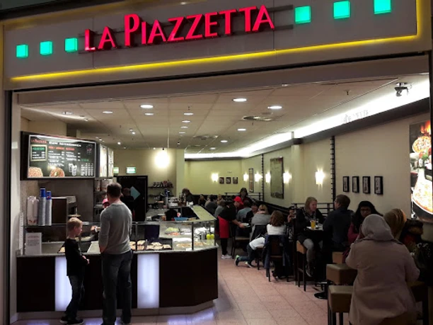 lapiazetta.jpg Das Bild zeigt den Imbiss "La Piazetta". Rechts sind Tische und Stühle zu sehen, während sich auf der linken Seite eine Theke befindet, an der ein Mann eine Bestellung aufgibt. Oberhalb prangt der Schriftzug "La Piazetta" in den Farben Grün und Rot.