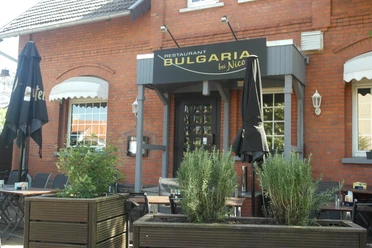 Bulgaria Das Bild zeigt die Außenansicht des Restaurants Bulgaria, das sich in einem roten Backsteingebäude befindet. Vor dem Eingang stehen geschlossenen schwarze Sonnenschirme, und über der Tür hängt ein schwarzes Schild mit dem goldenen Schriftzug "Bulgaria".