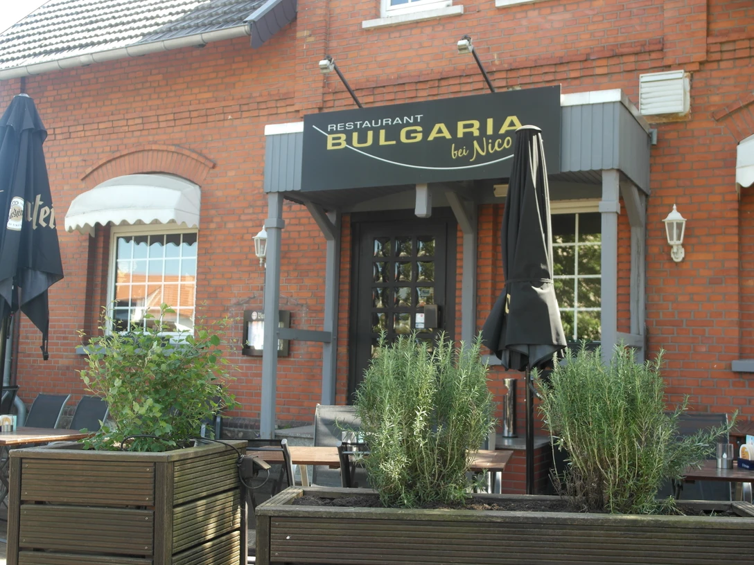 Bulgaria Das Bild zeigt die Außenansicht des Restaurants Bulgaria, das sich in einem roten Backsteingebäude befindet. Vor dem Eingang stehen geschlossenen schwarze Sonnenschirme, und über der Tür hängt ein schwarzes Schild mit dem goldenen Schriftzug "Bulgaria".
