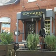 Bulgaria Das Bild zeigt die Außenansicht des Restaurants Bulgaria, das sich in einem roten Backsteingebäude befindet. Vor dem Eingang stehen geschlossenen schwarze Sonnenschirme, und über der Tür hängt ein schwarzes Schild mit dem goldenen Schriftzug "Bulgaria".