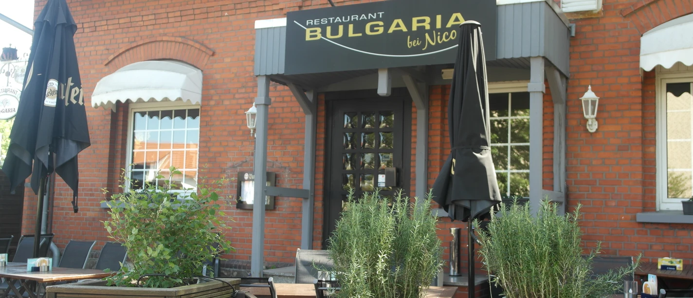 Bulgaria Das Bild zeigt die Außenansicht des Restaurants Bulgaria, das sich in einem roten Backsteingebäude befindet. Vor dem Eingang stehen geschlossenen schwarze Sonnenschirme, und über der Tür hängt ein schwarzes Schild mit dem goldenen Schriftzug "Bulgaria".