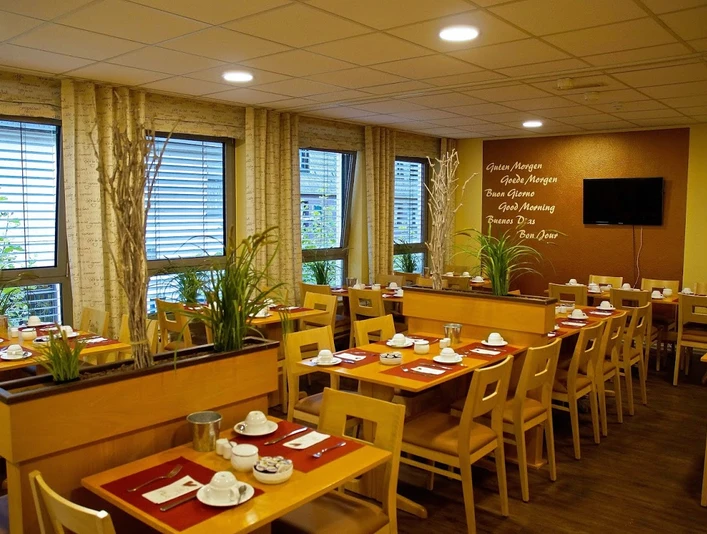 Frühstücksraum im Hotel Alberga in Mettmann Heller Frühstücksraum mit Holzmöbeln, eingedeckt für Gäste, bietet Ausblick durch große Fenster.