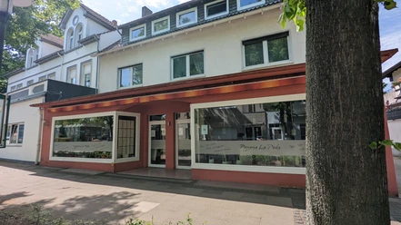 Pizzeria La Perla.jpg Zu sehen ist die Außenansicht der Pizzeria, deren Fassade in Rotbraun und Weiß gehalten ist. Die großen Fenster laden zu einem gemütlichen Mittagessen ein.