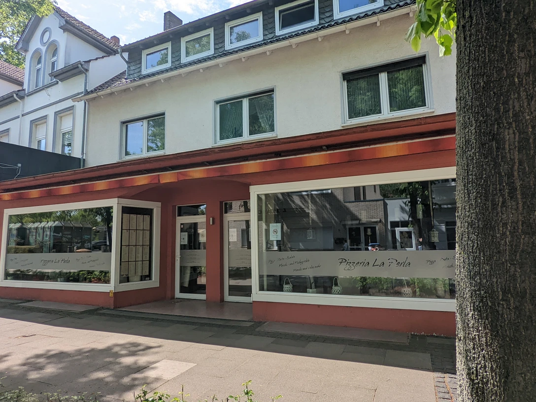 Pizzeria La Perla.jpg Zu sehen ist die Außenansicht der Pizzeria, deren Fassade in Rotbraun und Weiß gehalten ist. Die großen Fenster laden zu einem gemütlichen Mittagessen ein.