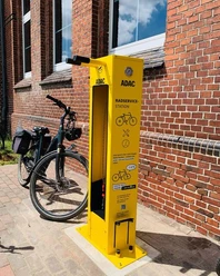 Fahrrad Reparaturstation.jpg Eine leuchtend gelbe Fahrrad-Reparaturstation von ADAC steht vor roten Backsteinwänden, mit Fahrrad ausgestattet.