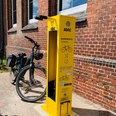 Fahrrad Reparaturstation.jpg Eine leuchtend gelbe Fahrrad-Reparaturstation von ADAC steht vor roten Backsteinwänden, mit Fahrrad ausgestattet.
