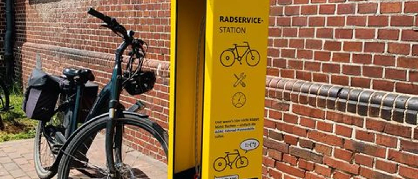Fahrrad Reparaturstation.jpg Eine leuchtend gelbe Fahrrad-Reparaturstation von ADAC steht vor roten Backsteinwänden, mit Fahrrad ausgestattet.