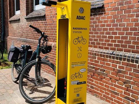 Fahrrad Reparaturstation.jpg Eine leuchtend gelbe Fahrrad-Reparaturstation von ADAC steht vor roten Backsteinwänden, mit Fahrrad ausgestattet.