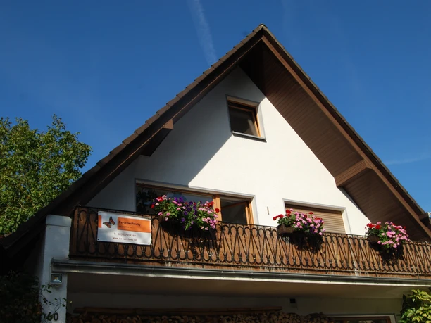 Außenansicht der Ferienwohnung Lang in Velbert Ferienwohnung mit Holzverzierungen und blühendem Balkon vor strahlend blauem Himmel in Velbert.