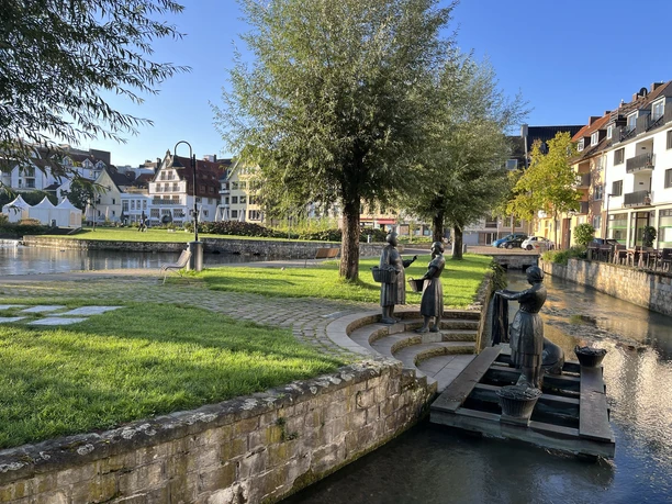 Drei bronzene Statuen stellen Waschfrauen an einem ruhigen Flussufer mit umliegenden Bäumen dar.