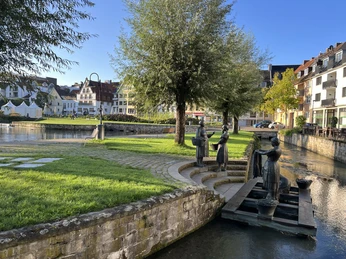 Waschfrauendenkmal an der warmen Pader Drei bronzene Statuen stellen Waschfrauen an einem ruhigen Flussufer mit umliegenden Bäumen dar.