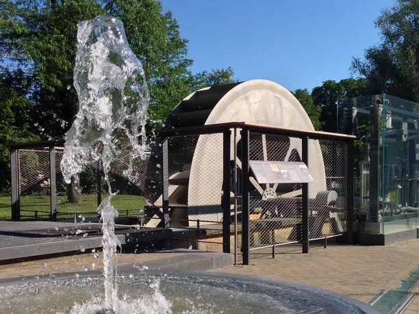 Ein historisches Wasserrad mit sprudelnder Fontäne im Vordergrund, umgeben von Grünflächen.