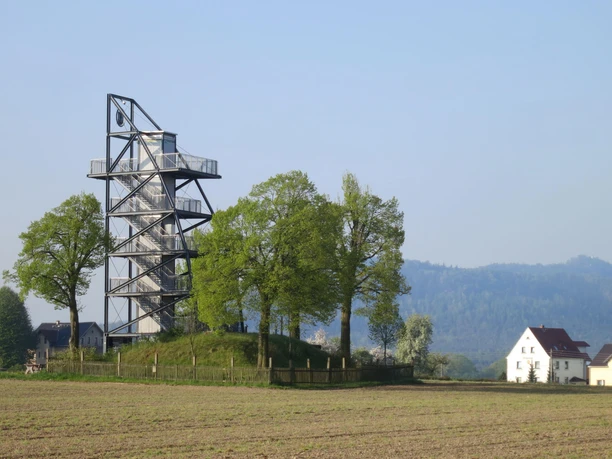 Aussichtsturm neben einem Feld mit zwei Häusern, bewaldete Hügel im Hintergrund.