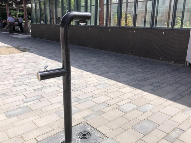 Ein moderner, minimalistischer Trinkwasserbrunnen aus Metall steht auf einem gepflasterten Bahnsteig.