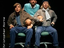 Drei Personen in einem humorvollen Theaterstück, sitzend auf Stühlen, mit expressiven Gesichtsausdrücken.