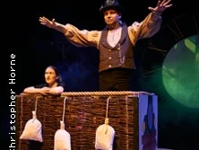 Zwei Personen in einer Theaterszene: Eine steht mit ausgebreiteten Armen, die andere lehnt an einem Korb.