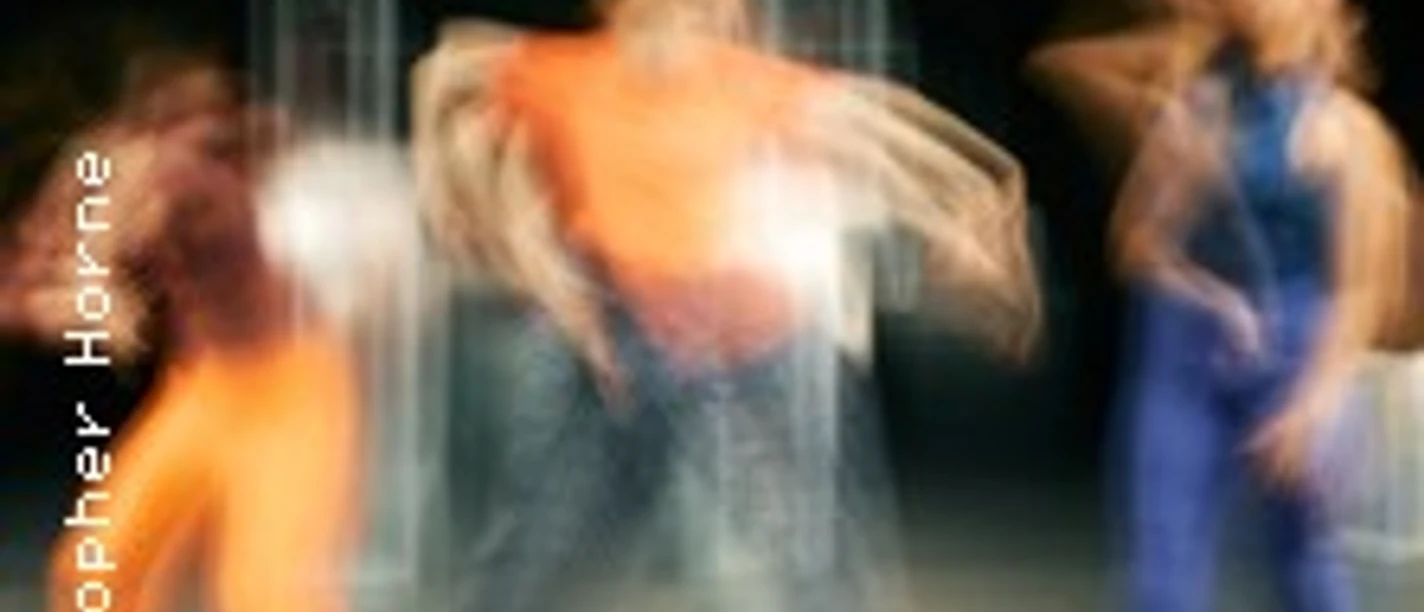 Drei Tänzer*innen bewegen sich dynamisch in verschwommenen, farbenfrohen Kostümen auf einer Bühne.